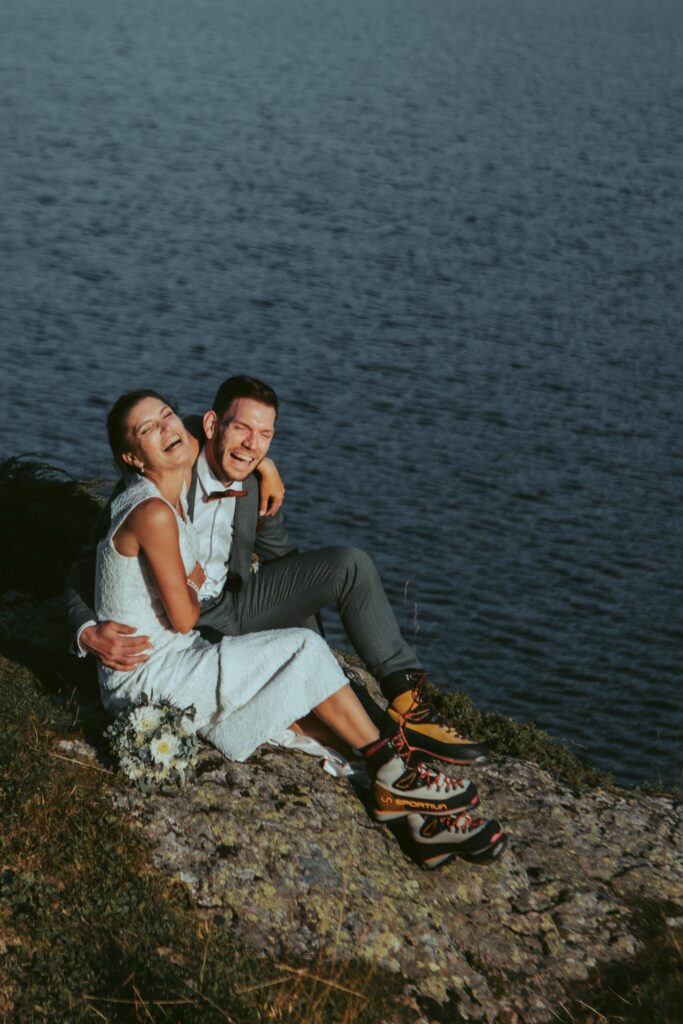 A couple sits toghether laughing on the rocks in front of a lake during their elopement in Switzerland