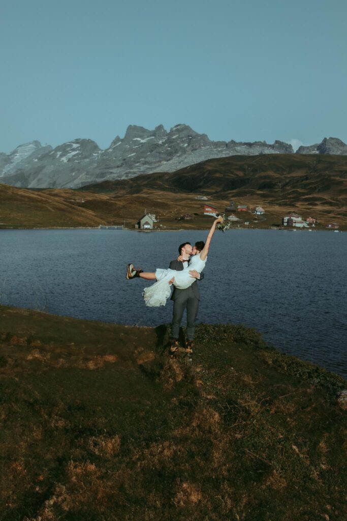 During their elopment in the Siwss Alps, a groom lifts up his bride and twirls her in front of an alpine lake, as she holds the flower bouquet in the air
