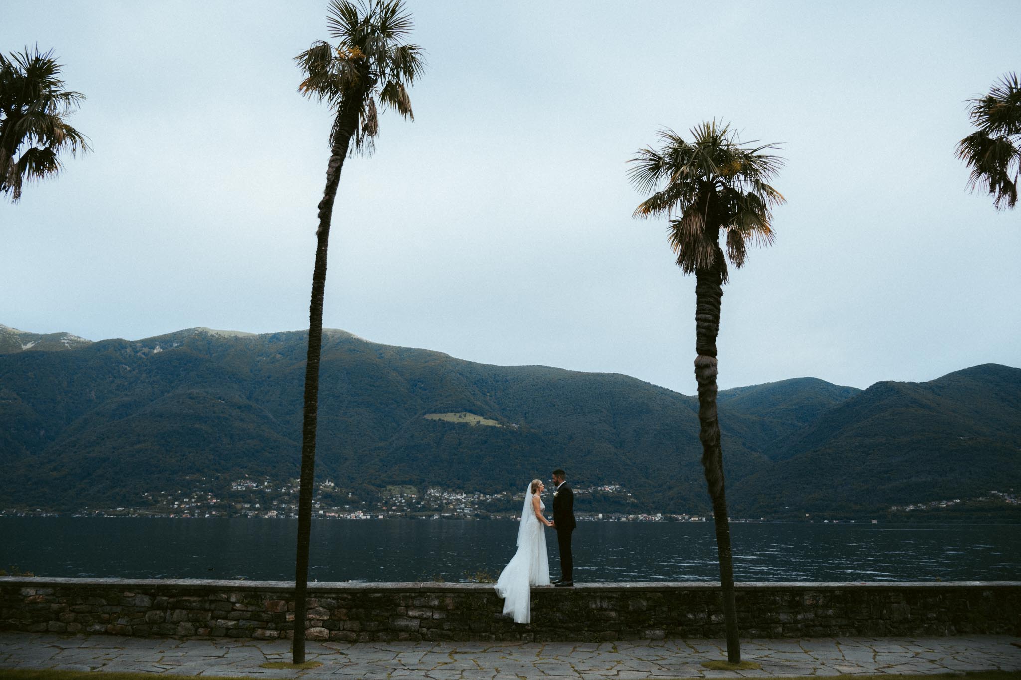 An eloping couple stands on a rockwall in front of the Ceresio lake and mountains in the distance after their wedding in Switzerland