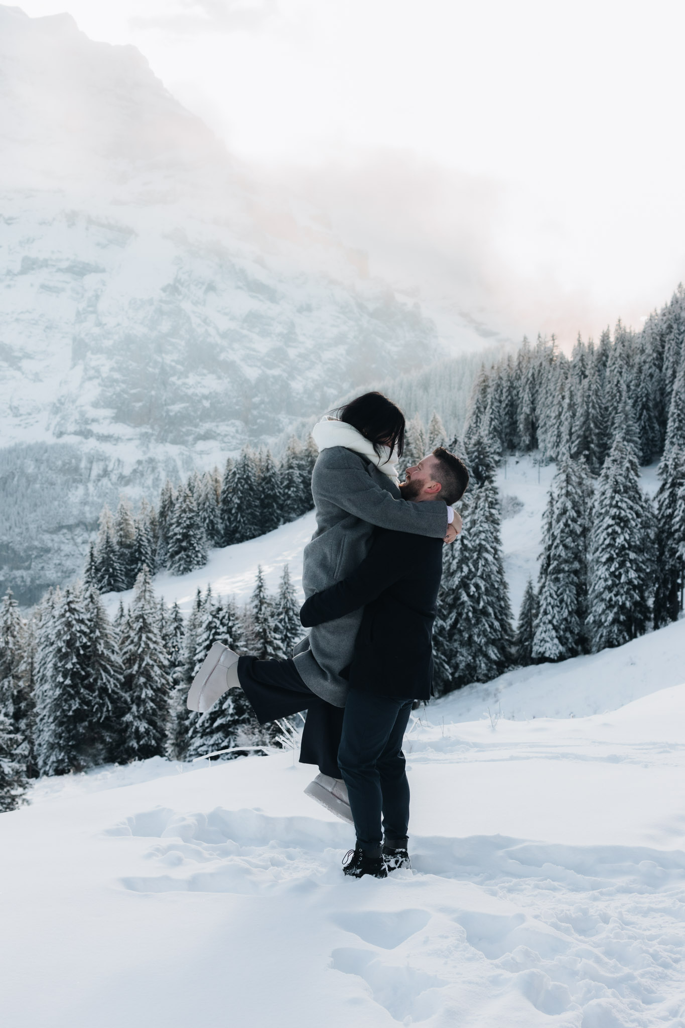 After a surprise proposal in the mountains of Lauterbrunnen, Switzerland, a man picks up his fiancée and twirls her around in front of snow-covered pinetrees and snow-capped mountain peaks 