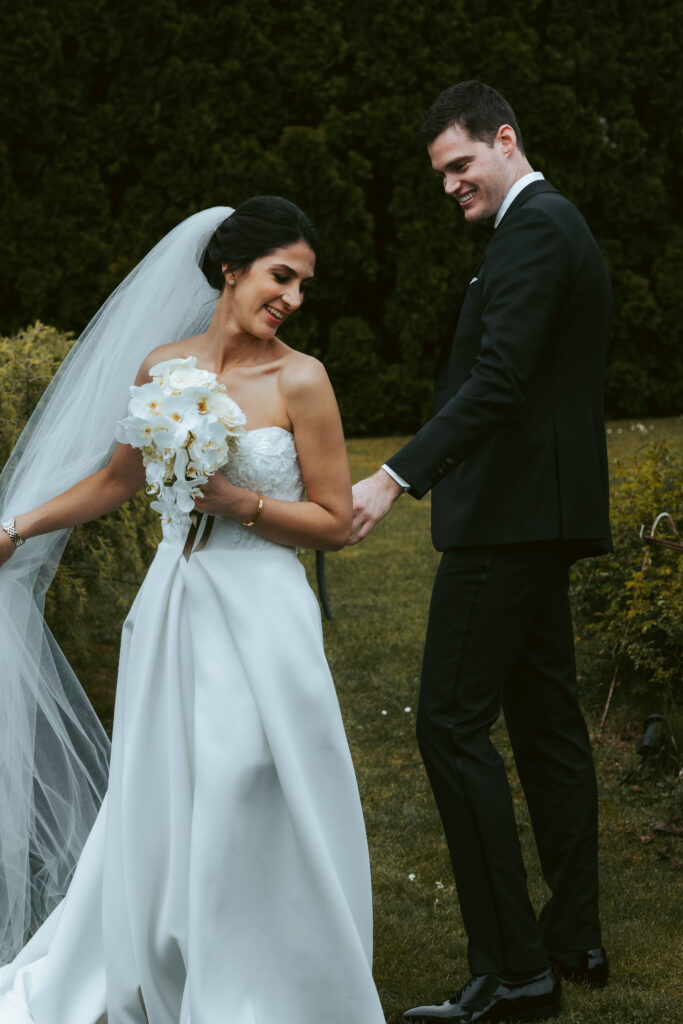 During their elopement in Switzerland on an overcast day, the bride spins around for the groom to see her wedding dress for the first time with a woodsy background, holding her veil and looking over her shoulder