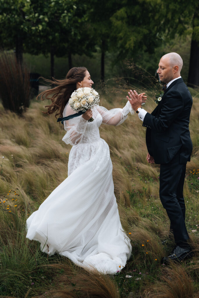 During their wedding in Switzerland a couple poses in a mountain wildflower field and holds hand while the wind blows the bride's hair and dress beautifuly in the wind