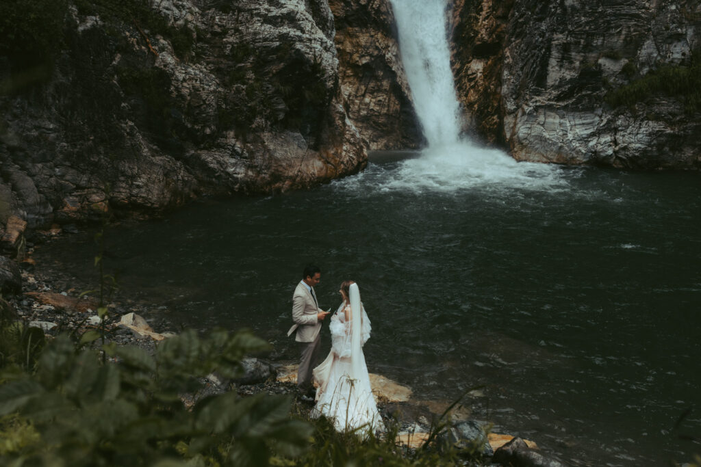 A couple stands on a small, rocky beach and reads their vows to each other in front of a waterfall during their elopement in Switzerland