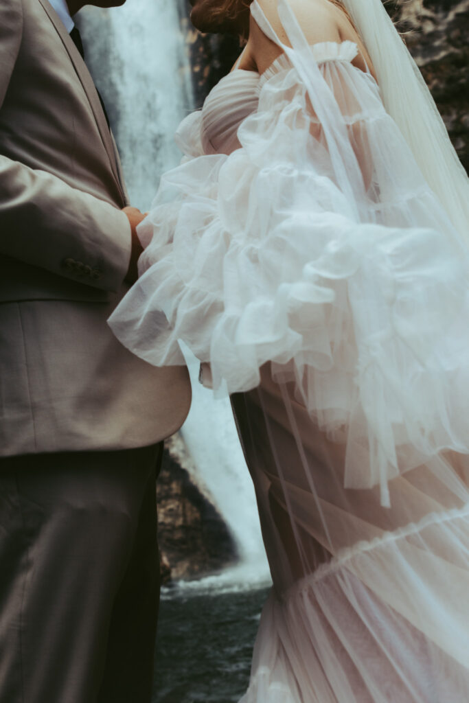 Close-up details of a bride and groom's wedding attire as they hold hands in front of a waterfall during their elopement in Switzerland
