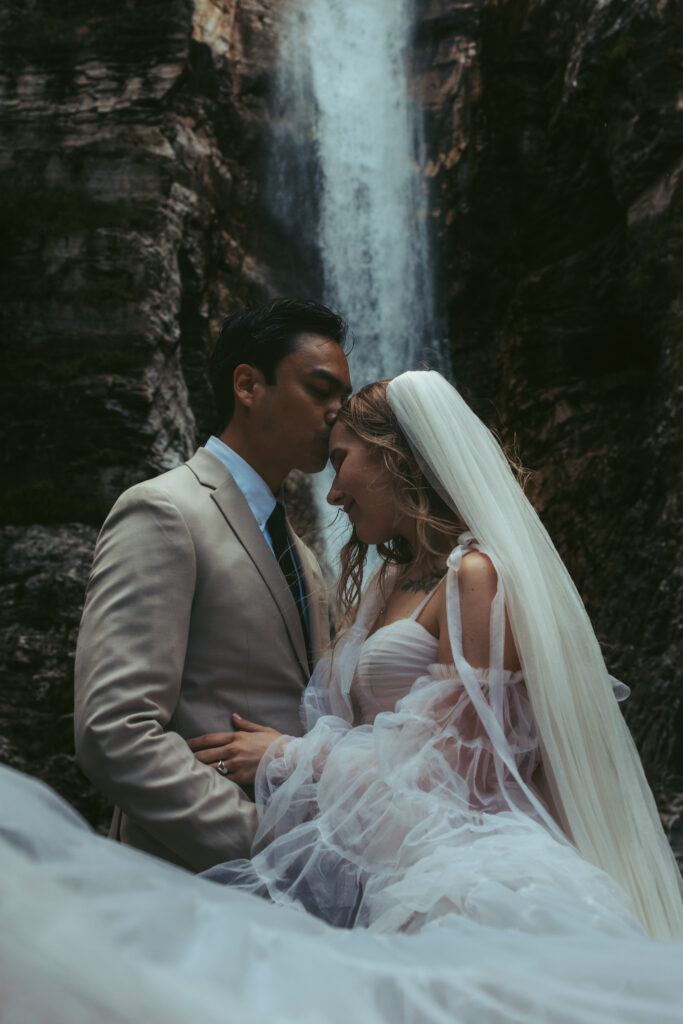 A groom kisses his bride's forehead during their elopement in front of a Swiss waterfall