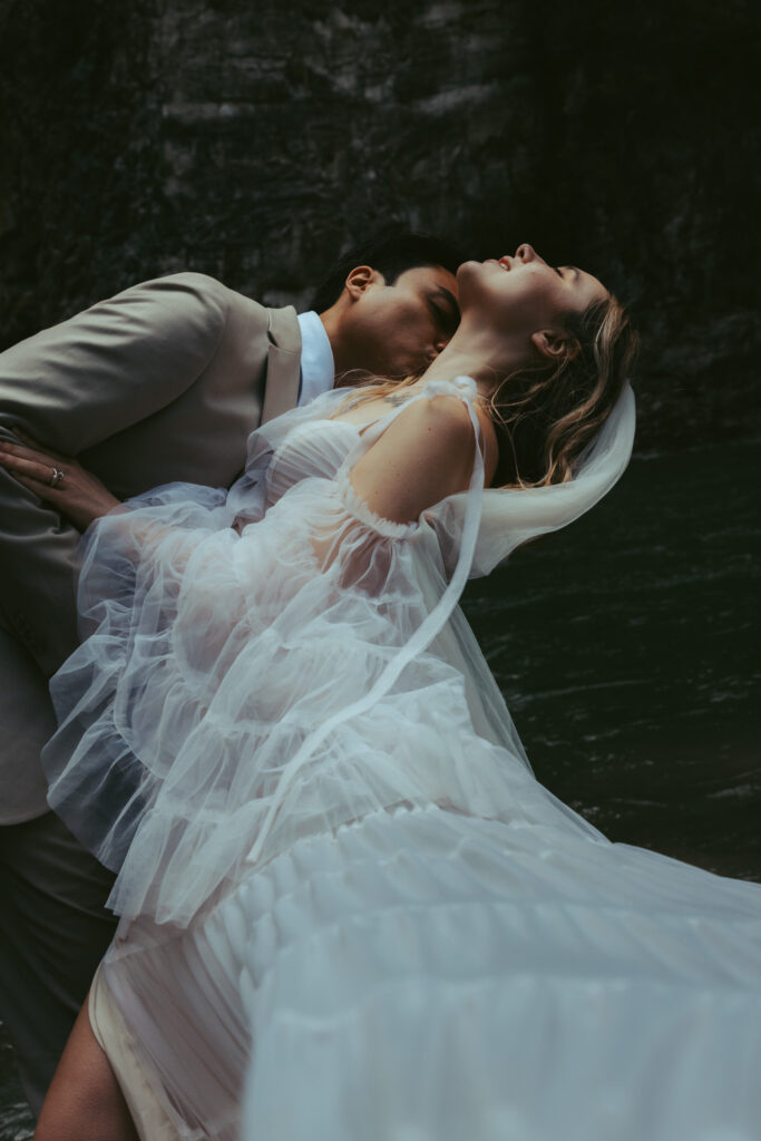 During their elopement in switzerland, a couple poses together in front of a waterfall: the groom leans over to kiss his wife on the neck, and she leans back with her body
