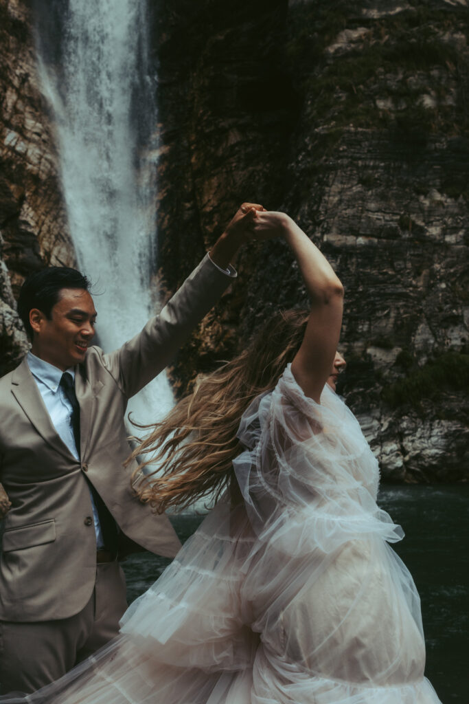 A groom twilrs his bride in front of a waterfall during their elopement in Switzerland, and her dress sways in the wind
