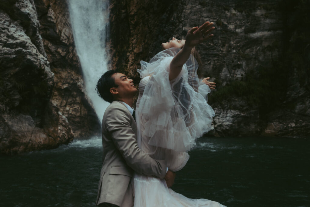 During their elopement in switzerland, a man lifts his bride into the air and twirls her in front of a mountain waterfall as her tulle sleeves nd veil blow in the wind