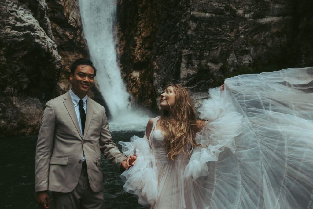 A couple elopes in front of a Swiss waterfall, the groom holding his bride's hand as she lifts her dress into the air with her other hand and looks at him lovingly