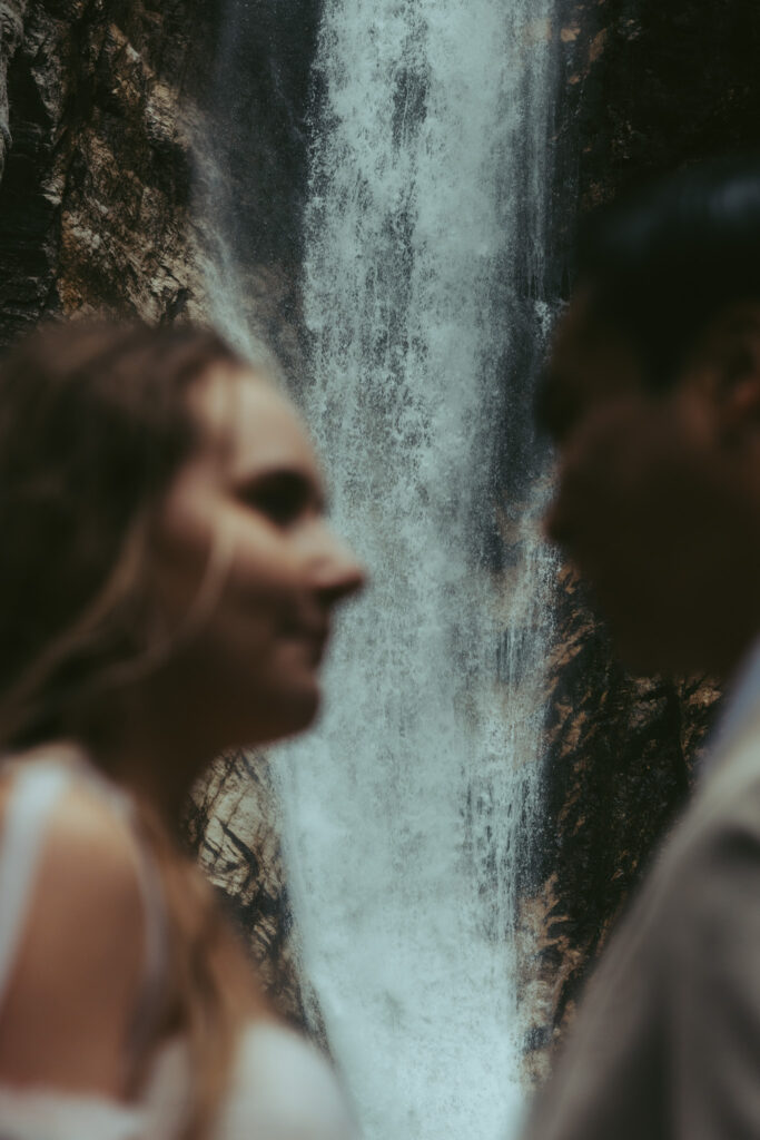 A couple stands in front of a waterfall during their elopement. Their faces are looking at each other but are blurred out, so the focus is on the waterfall behind them