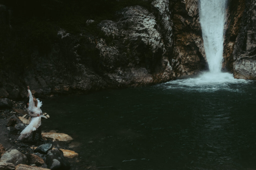 A couple stands on the side of a waterfall during their elopement in switzerland. The groom has picked up his bride and is twirling her around while her dress sways in the wind and she lifts her arm in the air