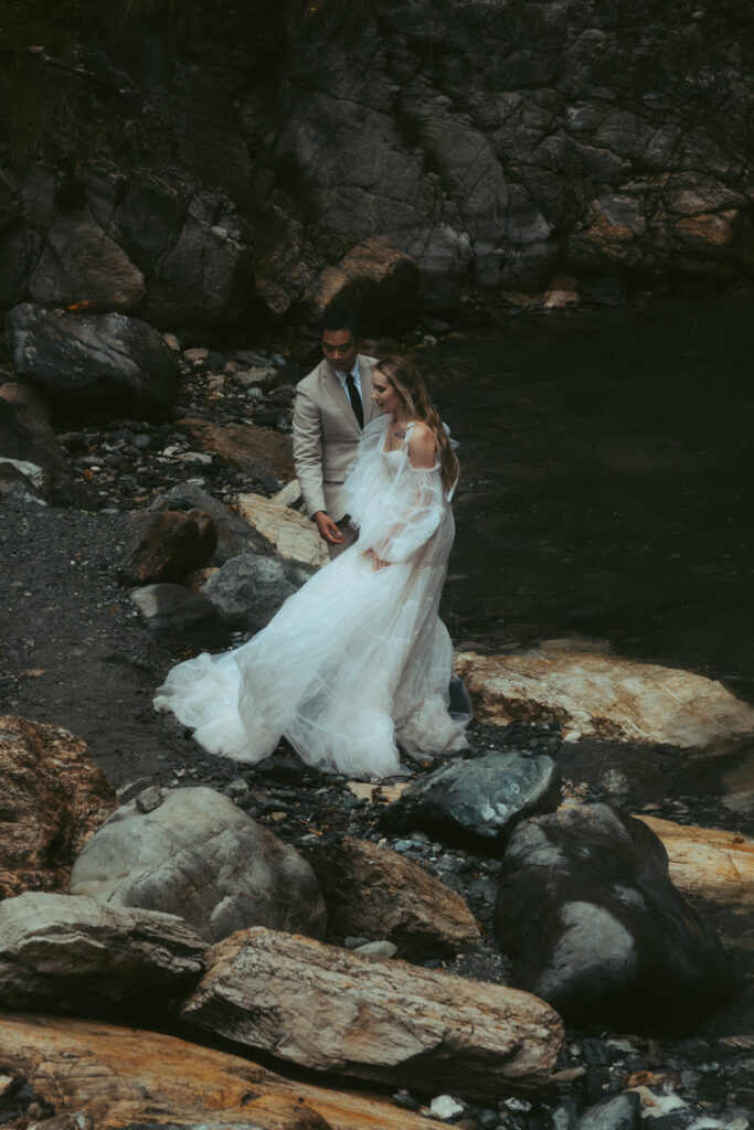 A couple elopes in front of a Swiss waterfall; standing on the rocky shore, the groom holds his bride from behind as she and her dress sway in the wind