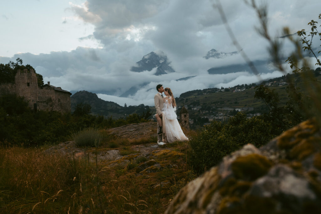A couple stands in the center of the image behind rocks and bushes, and leans into a kiss during their Swiss elopement
