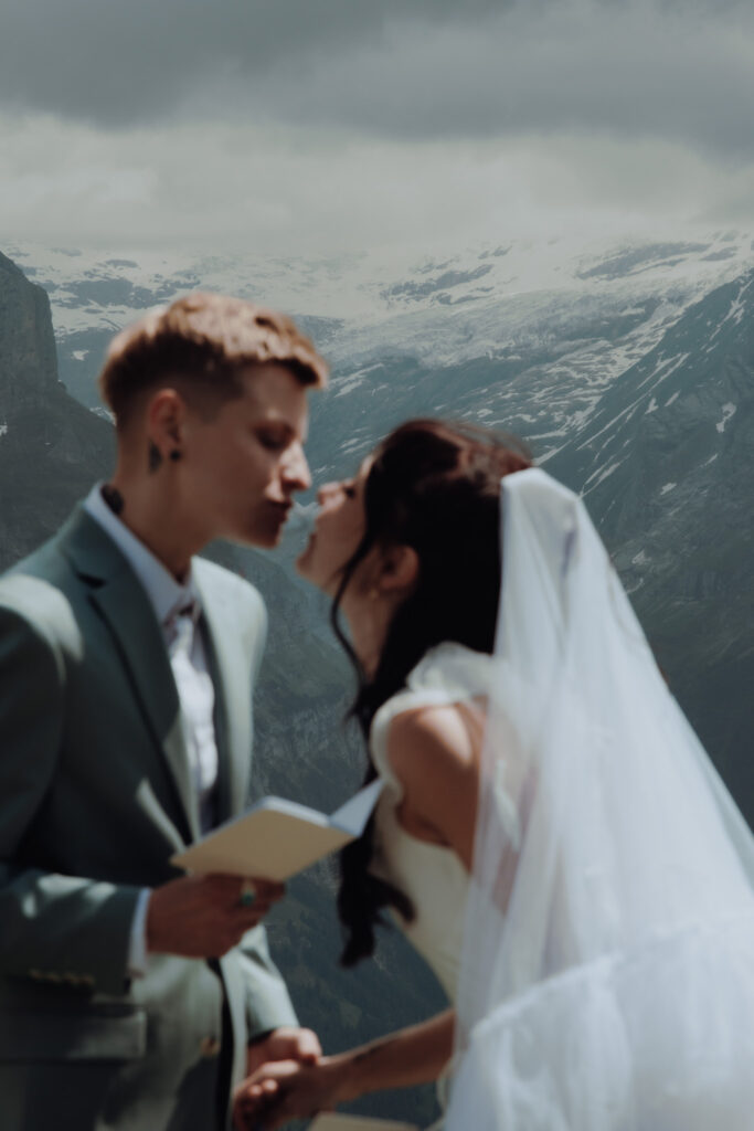A bride leans forward in the middle of the swiss alps to kiss her bride during their elopement, with the mountains in focus in the background
