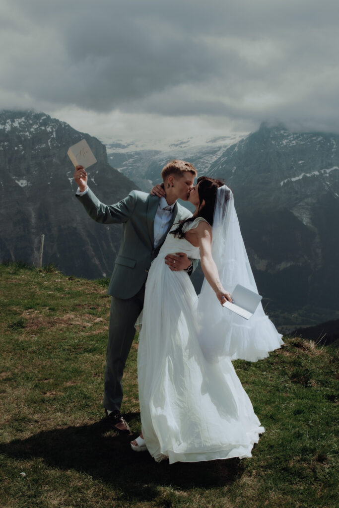 A couple elopes in the swiss alps. After having said their vows to each other, the couple kisses and raises their vow books in the air, and a glacier can be seen in the mountains behind them