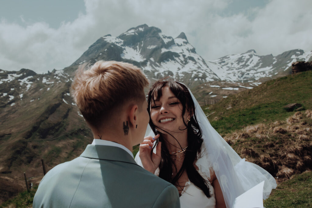 An eloping couple says their vows to each other in the middle of swiss mountain peaks. One bride looks at the other smiling, as her veil blows in the wind and she moves a strand of hair from her face