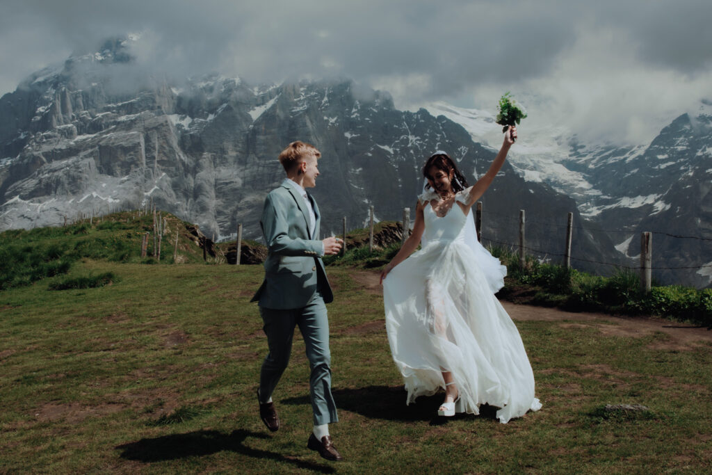 An eloping couple jumps in the air in front of the swiss alps, one woman holding a bouquet and the other woman holding her vow booklet as mountains and a glacier can be seen in the distance