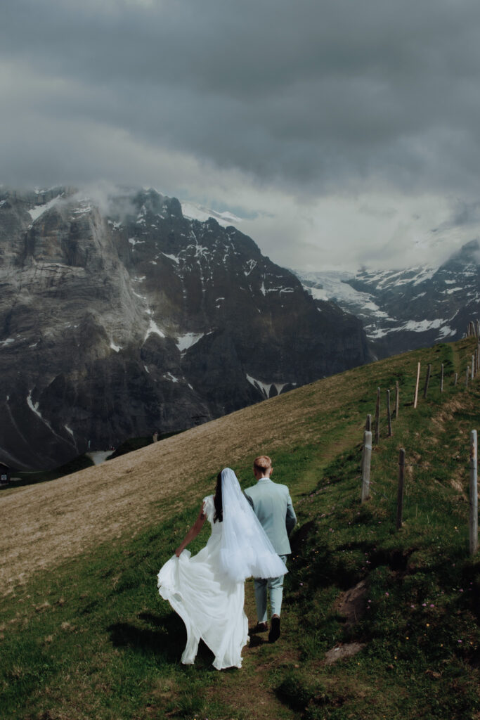 An eloping couple walks along a Swiss moutain ridge. One woman walks ahead and is holding the other woman's hand as they walk together away from the photographer