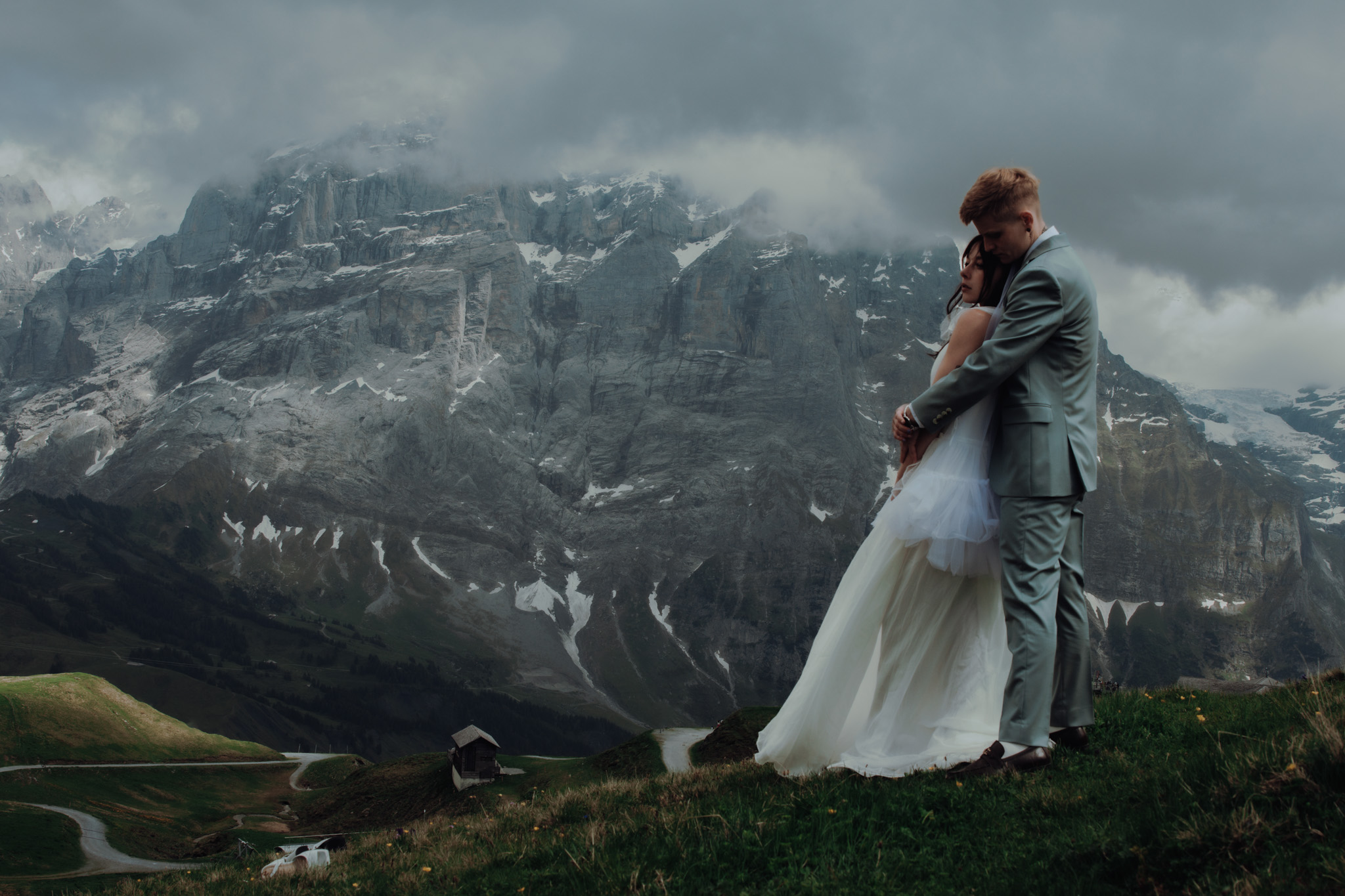 During their elopement in Grindelwald (Switzerland), two women hold each other, as one woman's wedding dress blows in the wind and mountain peaks can be seen behind them