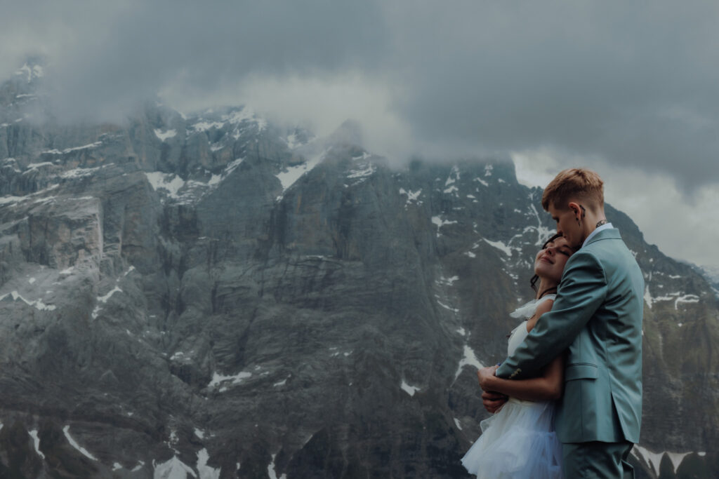 A couple elopes in the swiss alps- One woman holds the other from behind, lightly kissing her forehead in front of swiss alpine peaks