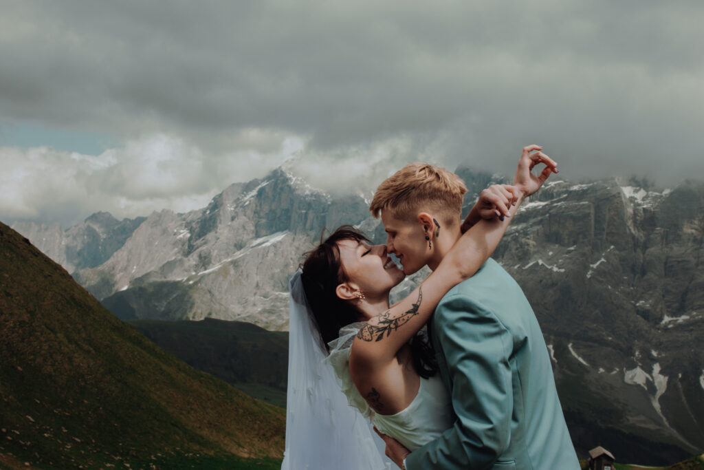 A couple leans into a kiss during their elopement in Grindelwald, Switzerland, as one woman's arms are wrapped around the other woman's wait, and mountain peaks can be seen behind them