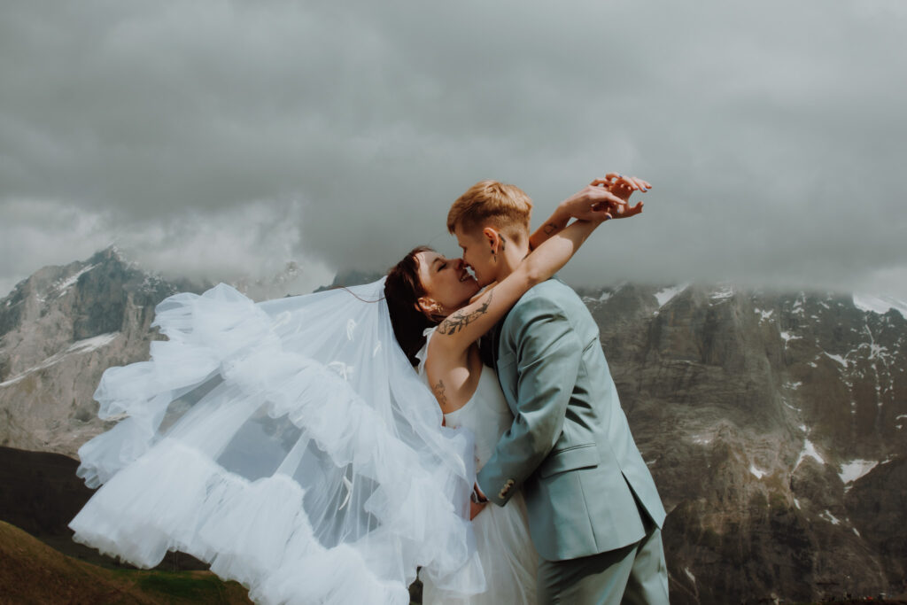 During their elopement in Grindelwald (Switzerland), a couple leans into a kiss during their elopement. One woman's veil blows in the wind while in the background there are swiss alpine peaks