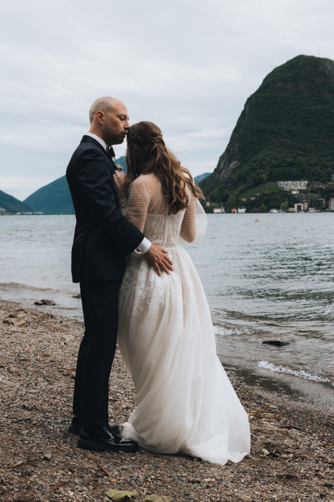 A married couple holds each other after their wedding in Switzerland, standing in front of a lake and mountains in the distance, while the groom kisses his bride's forehead