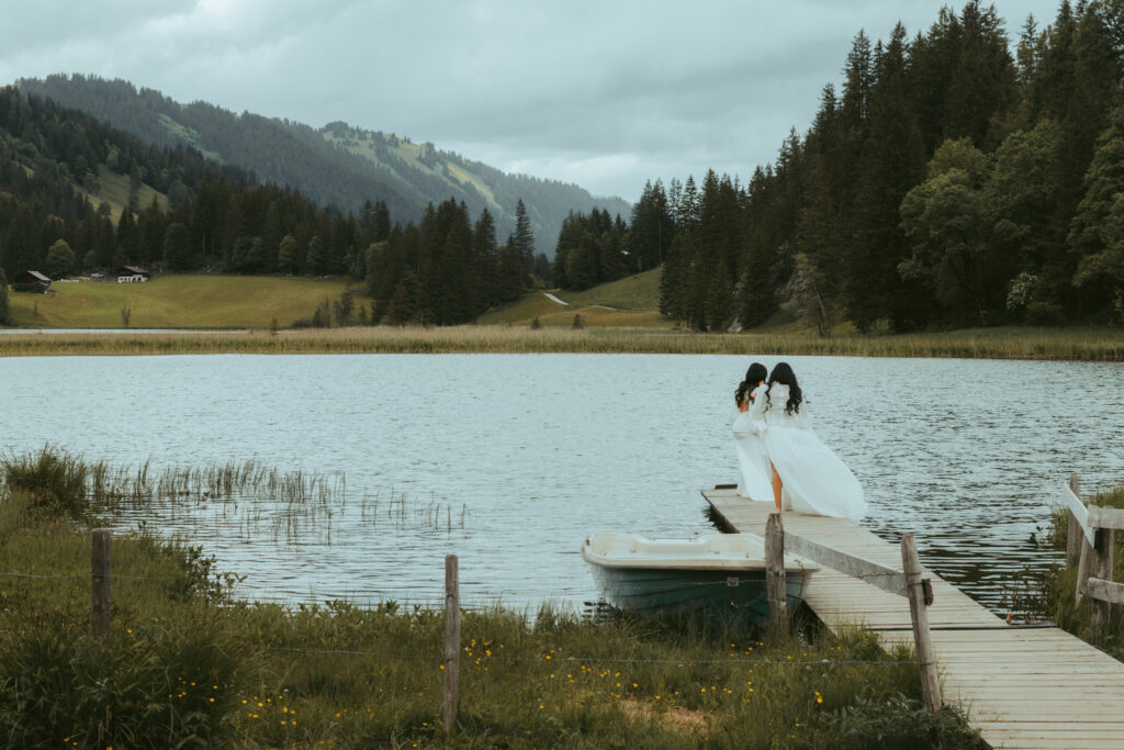 During their elopement in the swiss alps, a couple stands on a bridge on an alpine lake, holding each other close and letting the wind blow their dresses in the air