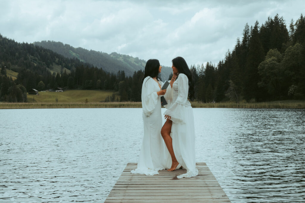 Switzerland elopement photographer immortalizes a couple as they elope in Switzerland on a lake surrounded by mountains. The two women stand and hold each other on a small wooden bridge overlooking the lake