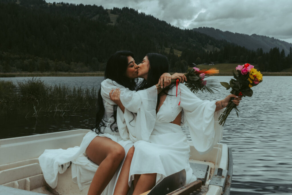 During their elopement in Switzerland, a couple sits on a boat and holds each other. Both women hold colorful flower bouquets in the air and smile while kissing