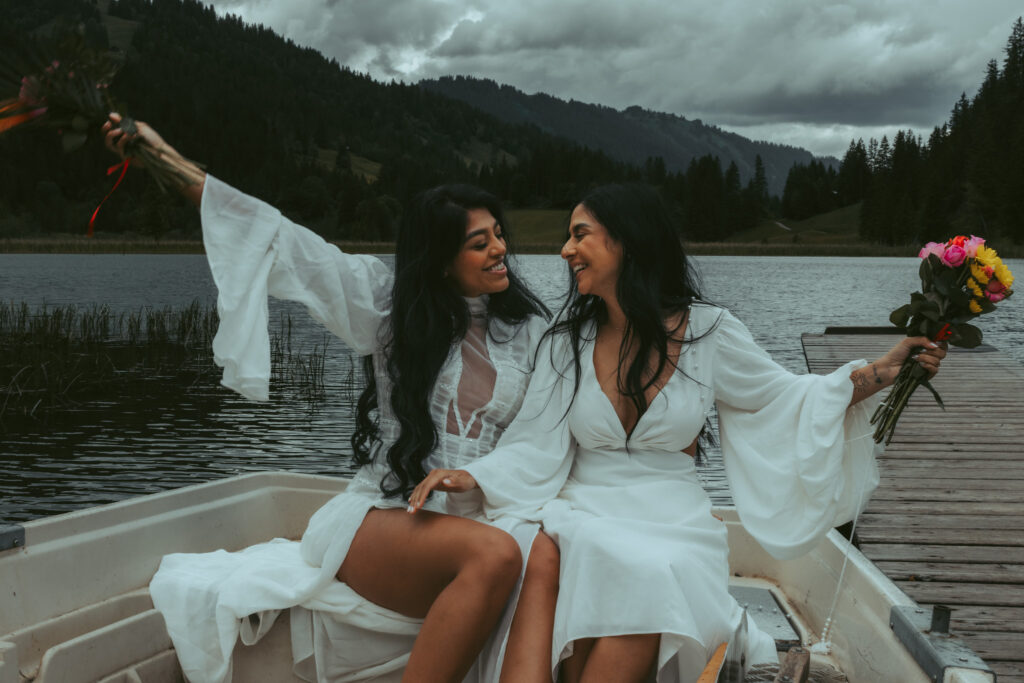 Two women celebrate their elopement in a boat on a swiss alpine lake. One woman raises her bouqet high in the air with her right hand as the other woman raises her bouquet on the other side, both smiling at each other
