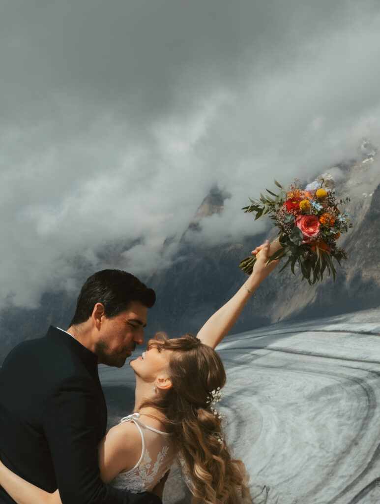 A bride and groom are about to kiss in front of a swiss glacier, as the bride holds her bouquet in the air