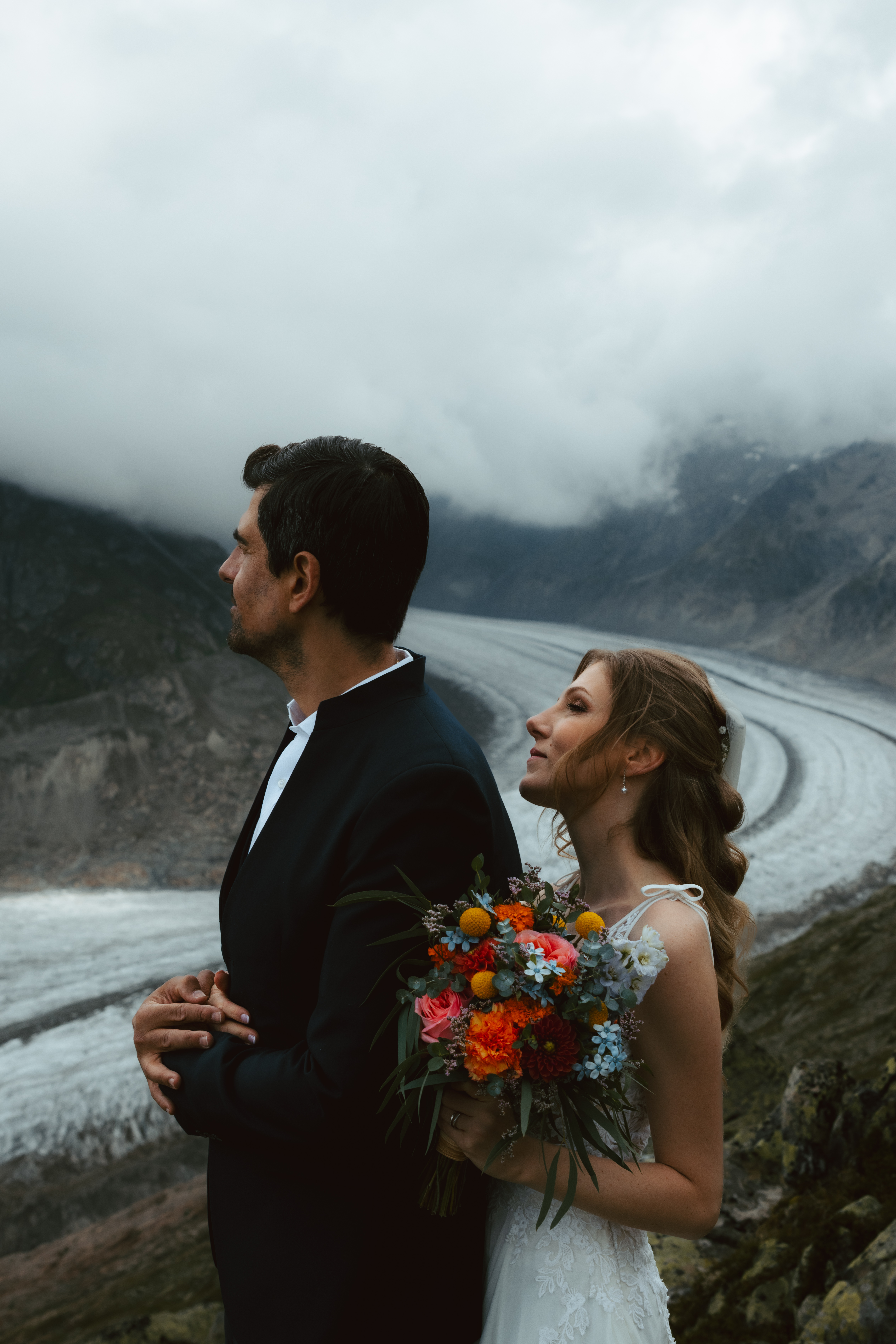 A bride holds her groom from behind during their glacier elopment in Switzerland, as her hair blows in the wind and her groom holds her hands on his chest
