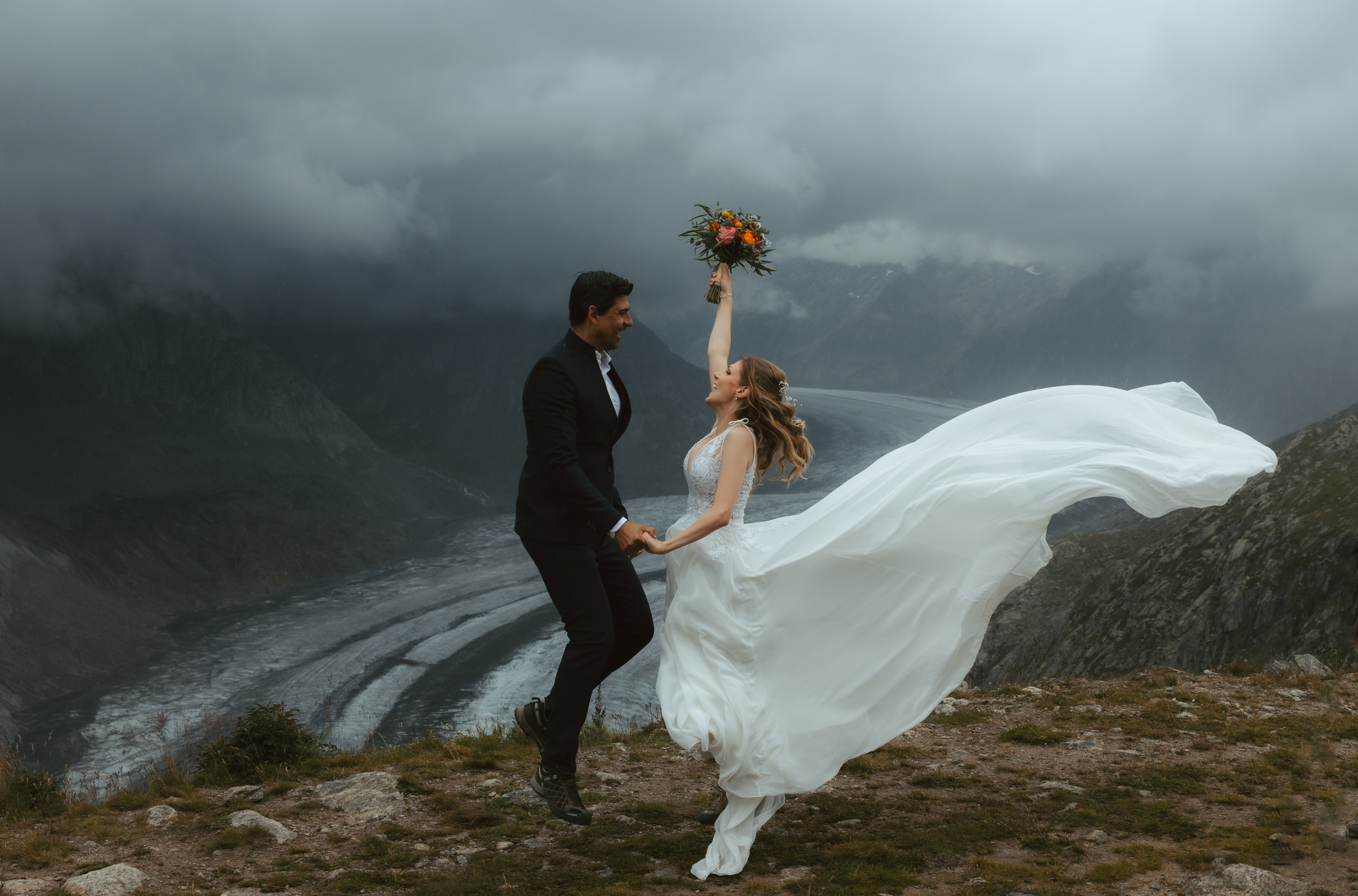 During their elopement at the Aletsch glacier, a couple jumps in the air - the bride's dress flows in the wind as she holds her flower bouquet in the air