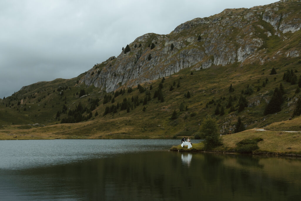 During their elopement wedding in Switzerland, a couple poses above a mountain lake, the bride twirling in her dress as the groom admires her