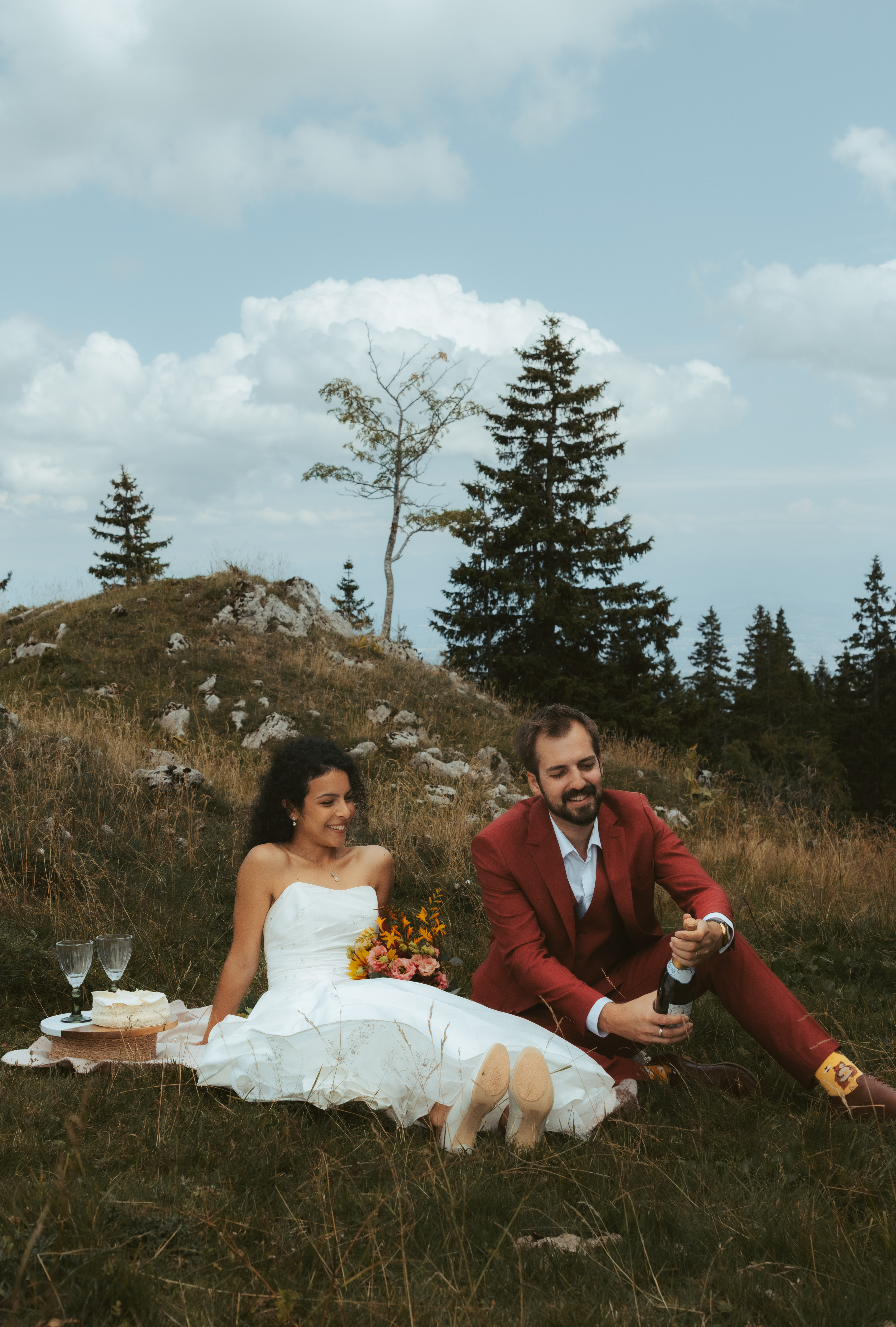 During their elopement in the swiss alps, a couple sits on a picnic, and the groom pops a bottle of champagne