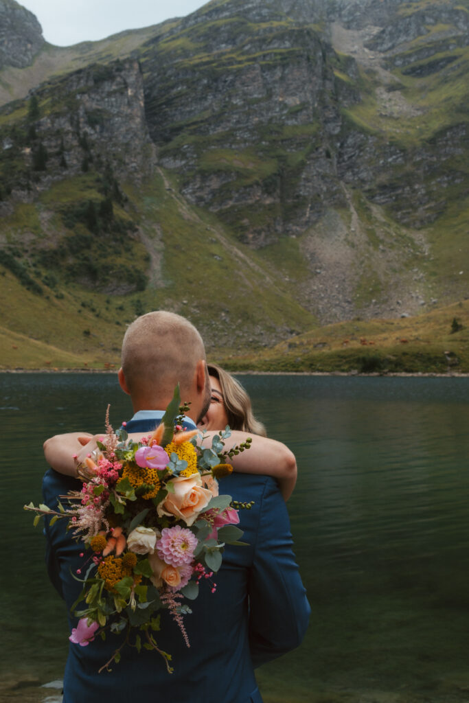 A couple hugs while standing in front of a mountain lake during their elopement in Switzerland. The bride is holding her bouquet with her arms wrapped around the groom's shoulders
