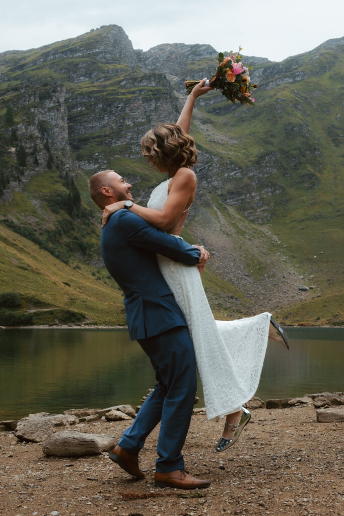 A groom lifts his bride in the air and spins her in front of a mountain lake in Switzerland during their elopement