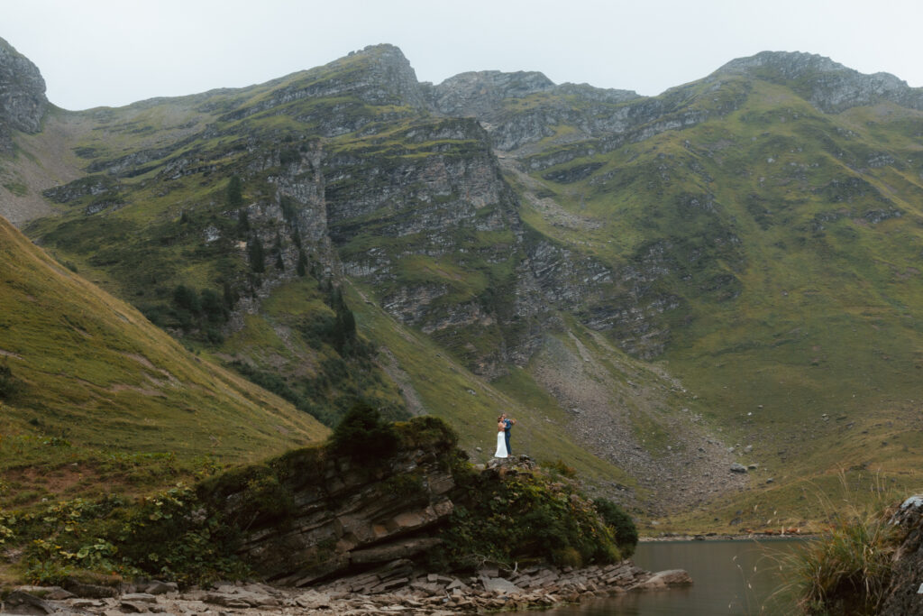 An eloping couple stands on a rock in the distance in front of massive Swiss peaks and a mountain lake