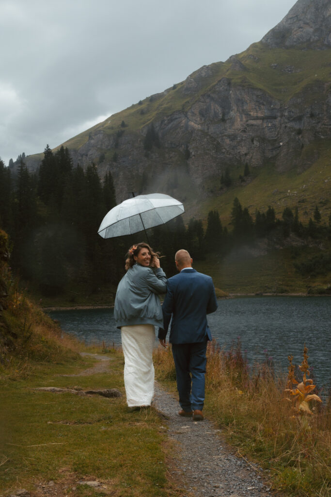 A couple walks on a mountain path in front of a lake, the bride holding an umbrella and looking back