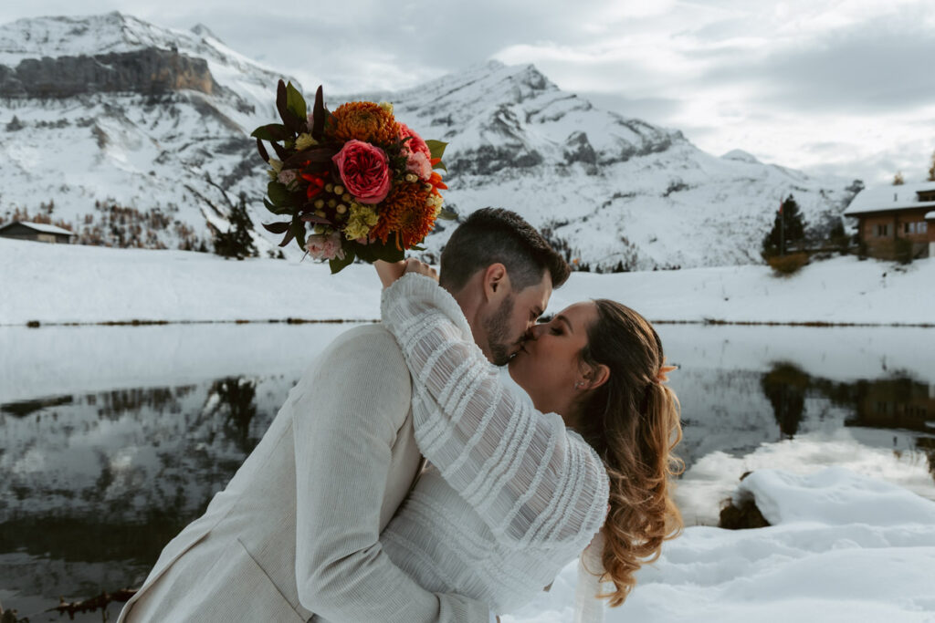 A couple leans into a kiss in front of a snow-covered alpine lake during their elopement in Switzerland