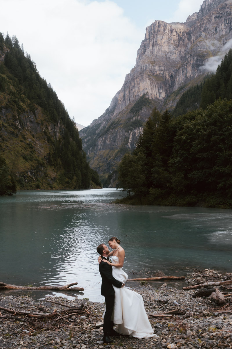 A couple leans into a kiss as the groom lifts his bride off the ground, all in front of the Swiss alps and a turqouise mountain lake