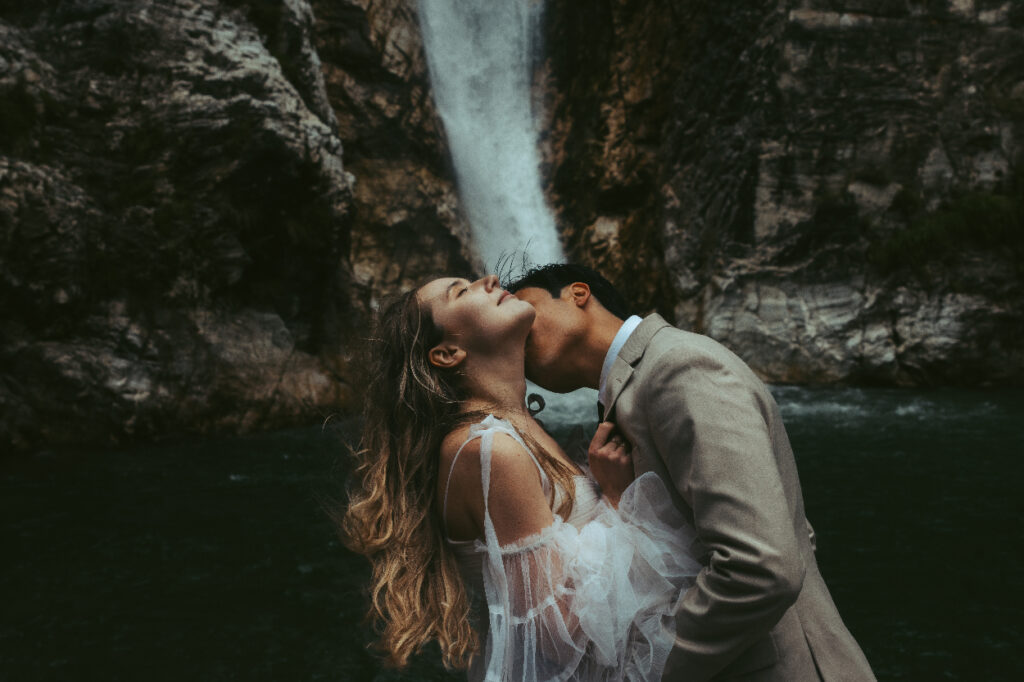 In front of a waterfall during their elopement in Switzerland, a man kisses his bride on the neck while she leans forward and her hair blows in the wind