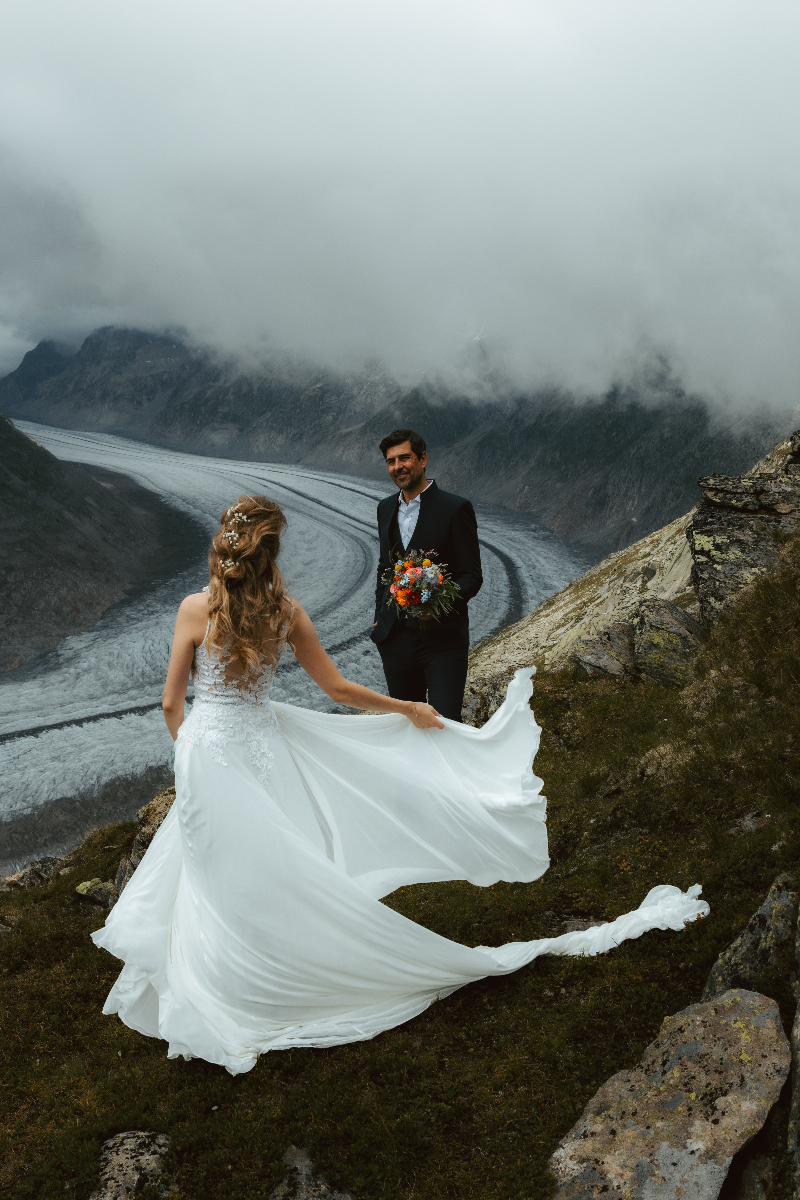 A bride twirls and lets her gown blow in the wind in front of her husband during their glacier elopement Swiss Alps