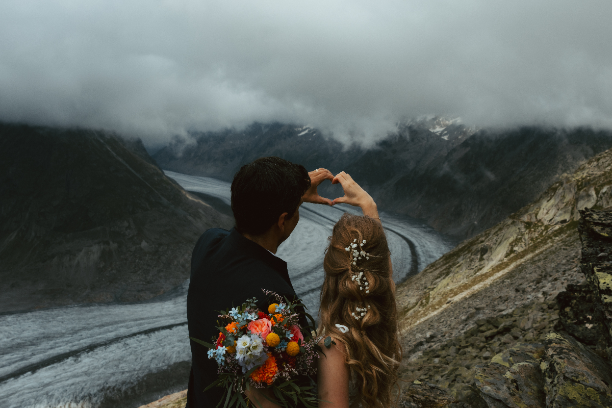 A couple makes a heart shape with their hands in front of a glacier during their elopement