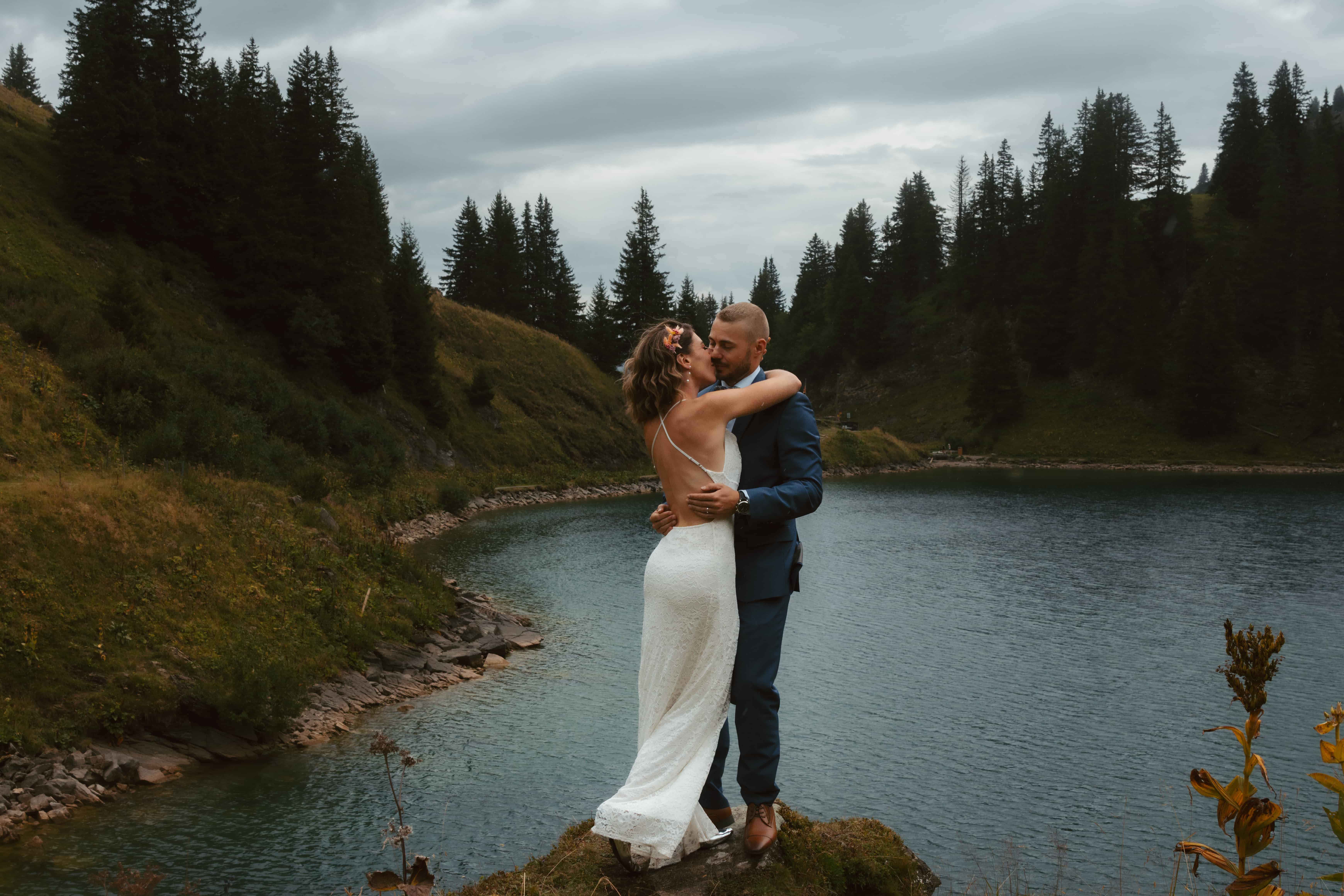 A couple holds each other close in the rain in front of an alpine lake and pine trees during their Swiss elopement